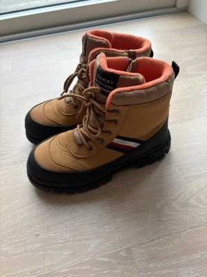 Bruna kängor från Tommy Hilfiger - Snygga bruna kängor från Tommy Hilfiger med orange fleecefoder, svart gummisula och klassiska snörningar. Kängorna har märkets logga på plösen och en randig detalj i rött, vitt och blått på sidan. Perfekta för kalla dagar och ger en sportig look.