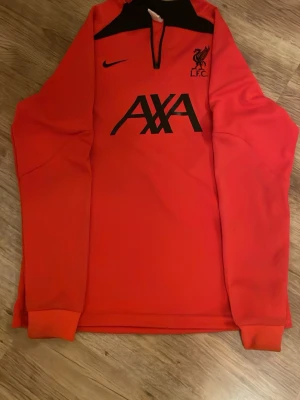 Liverpool Nike träningsjacka röd - Säljer en röd Liverpool FC träningströja från Nike med svart AXA-logga och klubbmärke på bröstet. Jackan har svart panel upptill och en praktisk half zip-dragkedja. Perfekt för dig som älskar fotboll och vill ha något snyggt att träna i.