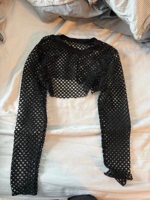 Svart croppad nät-topp - Säljer en svart croppad nät-topp med lång ärm. Toppen har ett coolt rutmönster och är perfekt för att styla över ett linne eller bh. Den har en tight passform och är riktigt edgy för dig som vill sticka ut.