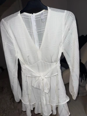 Vit omlottklänning med knytband - Snygg vit kortklänning med omlottdesign och knytband i midjan. Klänningen har v-ringning, långa ärmar och volangdetaljer nedtill. Tunt rutigt mönster i tyget ger en extra touch. Perfekt för studenten!