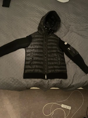 Svart dunväst med huva från Moncler - Snygg svart dunväst från Moncler med huva och dragkedja framtill. Västen har quiltad framsida, två fickor med dragkedja och stickade svarta ärmar. Klassisk Moncler-logga på ena ärmen. Perfekt för lager-på-lager och streetstyle.