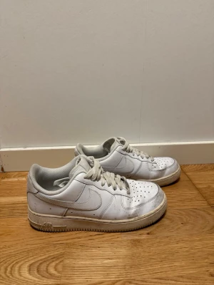 Vita Nike Air Force 1 låga sneakers - Klassiska vita Nike Air Force 1 sneakers i lågt utförande. Skorna har en stilren design med perforerad tå, vita skosnören och diskret Nike-logga på sidan. Tillverkade i läder med gummisula för bra grepp. Perfekta för en clean och tidlös look.
