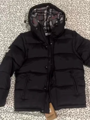 Svart pufferjacka från Burberry - Säljer en svart pufferjacka från Burberry med klassiskt rutigt innerfoder och huva. Jackan har dragkedja och knappar framtill samt två fickor med dragkedja. Perfekt för kalla dagar och riktigt snygg streetstil.