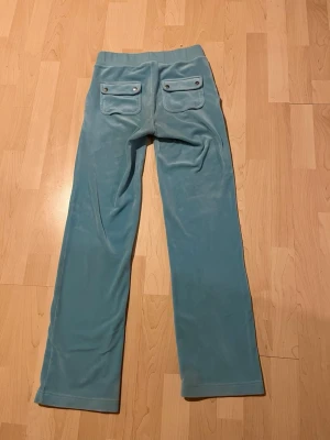 Blå velour mjukisbyxor Juicy Couture - Blåa mjukisbyxor i velour från Juicy Couture med raka ben, dragsko i midjan och stora fickor både fram och bak. Klassisk logga broderad på ena fickan framtill. Perfekta för en chill och trendig look. ✨🌊🐬