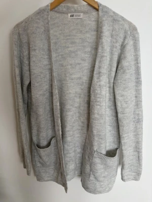 Ljusgrå kofta från H&M - Mysig ljusgrå kofta från H&M med öppen front och två praktiska fickor framtill. Koftan är långärmad och har en mjuk, stickad känsla som passar perfekt för lager på lager. Enkel och stilren design som funkar till det mesta.