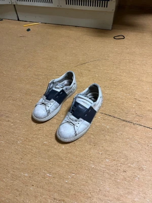 Valentino Garavani vita sneakers - Snygga vita sneakers från Valentino Garavani med bred mörkblå läderdetalj över sidan och klassisk snörning. Skorna har en låg profil och är tillverkade i läder med logotyp på plösen. Perfekta för dig som gillar exklusiv streetstyle.