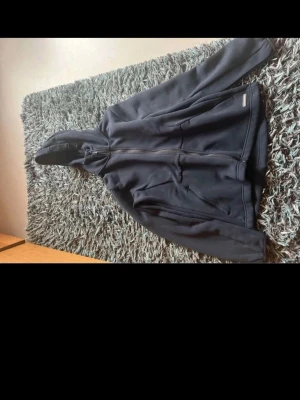 Blå hoodie från Cutter & Buck - Snygg blå hoodie från Cutter & Buck med dragkedja framtill och två fickor. Tillverkad i CB DryTec-material som andas och känns skönt mot huden. Klassisk passform med huva och diskret logga på bröstet. Perfekt för chill eller träning.