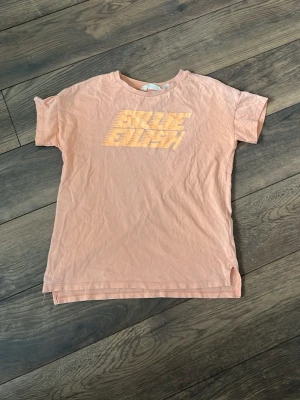 Ljusorange kortärmad t-shirt H&M - T-shirt i ljusorange från H&M med trycket 'BILLIE EILISH' framtill i gul ton. Klassisk rund hals och rak passform. Tillverkad i mjuk bomull som känns skön mot huden. Perfekt för dig som gillar en avslappnad och trendig stil.