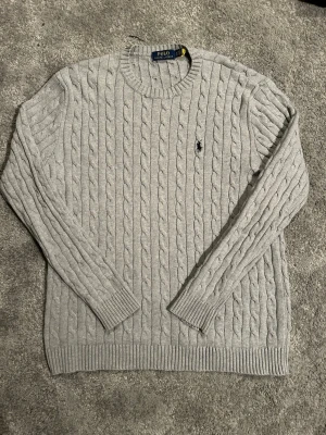 Grå kabelstickad tröja Polo Ralph Lauren - Snygg grå kabelstickad tröja från Polo Ralph Lauren med klassisk rund halsringning och broderad logga på bröstet. Tillverkad i mjuk bomull och har ribbade muddar vid ärmslut och nederkant. Perfekt för en stilren och avslappnad look.