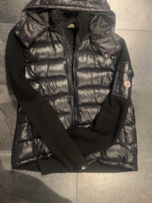 Moncler cardigan - Snygg svart dunväst från Moncler med glansig finish, stickade svarta ärmar och avtagbar huva. Västen har dragkedja framtill och Moncler-logga på ena ärmen. Perfekt för lager-på-lager. Storlek XL men sitter mer som L