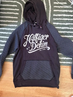 Svart Hilfiger Denim hoodie - Snygg svart hoodie från Hilfiger Denim med stor vit logga på bröstet och quiltade detaljer på ärmar och ficka. Tröjan har huva med dragsko och en stor magficka. Perfekt för en avslappnad och cool stil.