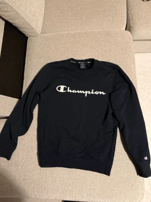 Svart sweatshirt från Champion - Säljer en svart sweatshirt från Champion med stort vitt logotryck över bröstet och liten logga på ärmen. Tröjan har rund halsringning och ribbade muddar vid ärmslut och nederkant. Perfekt för en avslappnad och sportig stil.