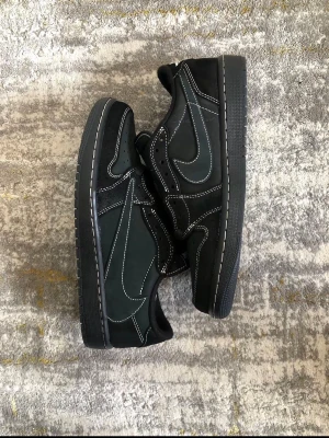 Svarta Nike Air Jordan 1 Low Travis Scott - Säljer ett par svarta Nike Air Jordan 1 Low Travis Scott sneakers med vita kontrastsömmar och omvänd Swoosh. Skorna har detaljer i mocka och läder samt broderade loggor på hälen. Extra röda skosnören medföljer. Perfekta för dig som gillar streetwear och unika samarbeten.