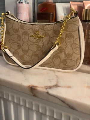 Beige axelremsväska från Coach - Snygg beige handväska från Coach med klassiskt monogrammönster och guldfärgade detaljer. Väskan har en vit kant, kedja och justerbar axelrem i vitt och guld. Perfekt storlek för det viktigaste och ger en trendig touch till din outfit.