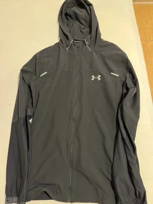 Svart vindjacka från Under Armour - Snygg svart vindjacka från Under Armour med hel dragkedja och huva. Jackan har reflekterande detaljer på bröstet och logga på vänster sida. Tillverkad i lätt och smidigt material som passar perfekt för aktiva dagar utomhus. Sitter som M och L 