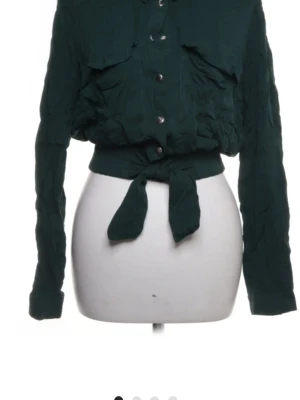 Mörkgrön knytblus med fickor - Snygg mörkgrön blus med knytband i midjan och långa ärmar. Tröjan har två bröstfickor med lock, knappar framtill och en lätt skrynklig textur. Perfekt för dig som vill ha en trendig och unik look.