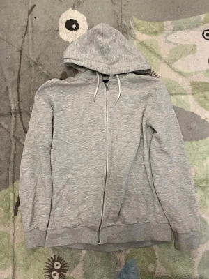 Ljusgrå zip hoodie med huva lager 157 - En stilren ljusgrå hoodie med dragkedja framtill och justerbar huva med vita snören. Tillverkad i mjukt bomullsmaterial och har ribbade muddar vid ärmar och nederkant. Perfekt för en avslappnad look och enkel att matcha med andra plagg.