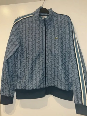 Blå mönstrad vindjacka och byxor från Lacoste  - Snygg blå vindjacka från Lacoste med monogrammönster och klassisk krokodillogga på bröstet. Jackan har dragkedja framtill, ribbade muddar och sportiga ränder längs ärmarna. Regular fit och perfekt för en streetwear-look. Storlek L på jackan, storlek XL på byxor men sitter som L. Använd 2-3 gånger.