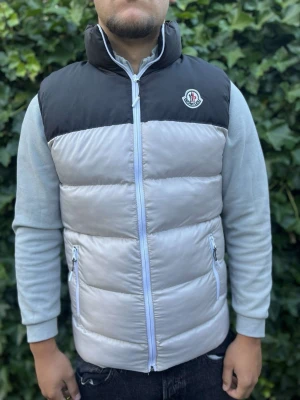 Moncler dunväst svart och grå - Snygg dunväst från Moncler i svart och ljusgrått med hög krage och dragkedja framtill. Västen har två fickor med dragkedja och Moncler-logga på bröstet. Perfekt för lager-på-lager och riktigt trendig look.