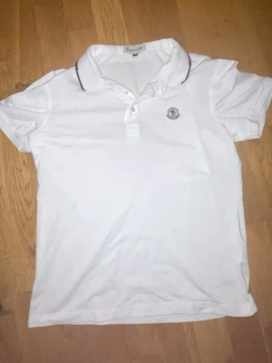 Vit pikétröja från Monc - Snygg vit pikétröja från Moncmed klassisk krage och korta ärmar. Tröjan har knäppning framtill och en liten Monc-logga på bröstet. Kragen har en tunn röd och blå rand som detalj. Perfekt för en stilren och sportig look.