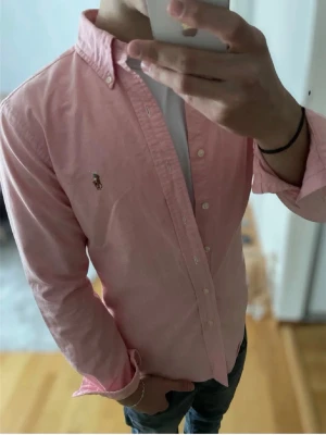 Rosa skjorta från Ralph Lauren - Klassisk rosa skjorta från Ralph Lauren med button-down krage och broderad logga på bröstet. Skjortan har långa ärmar, knappar framtill och är tillverkad i mjuk bomull. Perfekt för en clean och stilren look.