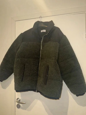 Grön teddyjacka från Peer One - Mysig teddyjacka i mörkgrönt med svarta detaljer från Peer One. Jackan har hög krage, dragkedja framtill och två fickor med dragkedja. Materialet är fluffigt teddytyg kombinerat med slätare tyg upptill. Perfekt för kalla dagar och riktigt snygg streetstil. Storlek XL sitter som L.