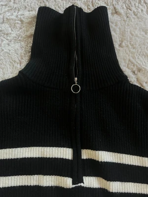 Svart stickad tröja med dragkedja - Svart ribbstickad tröja med hög krage och silverfärgad dragkedja med ringdetalj. Två vita horisontella ränder nedtill ger en sportig look. Perfekt för lager på lager och streetstyle. Köpt på Zalando om jag minns rätt.