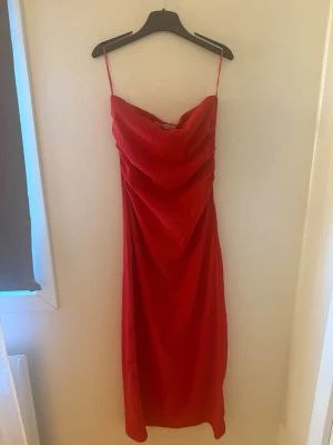 Röd draperad midiklänning - Säljer en röd midiklänning helt ny och ej använd med tunna axelband och draperad front. Klänningen har en mjuk, något glansig textur och faller snyggt längs kroppen. Perfekt för dig som vill sticka ut med en klassisk och trendig look.