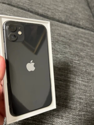  iPhone 11 med dubbla kameror - Snygg svart iPhone 11 med stilren glasbaksida och rundade kanter. Telefonen har dubbla kameror på baksidan och en stor skärm med tunn ram. Klassisk Apple-design med loggan centrerad på baksidan. Perfekt för dig som gillar modern teknik och enkel användning.