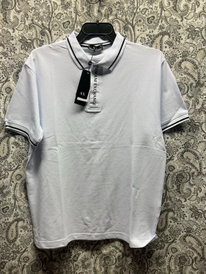 Vit piké från Armani Exchange - Klassisk vit pikétröja från Armani Exchange med svarta detaljer på krage och ärmslut. Kortärmad modell med rak passform och mjukt bomullsmaterial. Perfekt till jeans eller shorts för en clean look.