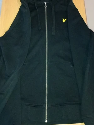 Svart zip hoodie från Lyle & Scott - Säljer en svart hoodie med dragkedja från Lyle & Scott. Klassisk design med huva, fickor framtill och det ikoniska gula fågelmärket broderat på bröstet. Perfekt för en chill och stilren look. Materialet känns mjukt och bekvämt.