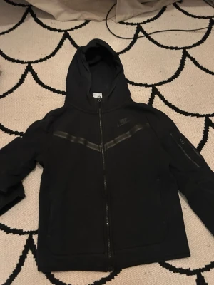 Svart hoodie från Nike med dragkedja - Säljer en svart hoodie från Nike med hel dragkedja framtill och huva. Hoodien har diskreta svarta detaljer över bröstet och en ficka med dragkedja på ena ärmen. Tillverkad i mjukt material, perfekt för en sportig och avslappnad stil.Sönder vid Nike märket lite grann men märks inte men därför är den så billig.