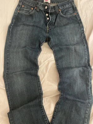 Levi's 501 blå jeans med knappar - Klassiska Levi's 501 jeans i blå tvätt med raka ben och knappgylf. Jeansen har fem fickor, kontrastsömmar och den ikoniska läderlappen bak i midjan. Perfekta för en avslappnad och tidlös stil.