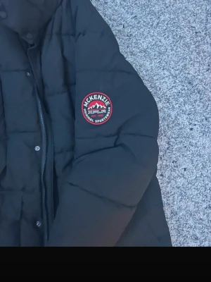 Svart dunjacka från McKenzie - Säljer en svart dunjacka från McKenzie med huva och patch på ärmen. Jackan har dragkedja och knappar framtill samt stora fickor. Perfekt för kalla vinterdagar och riktigt skön att bära. Klassisk, stilren design som passar till det mesta.