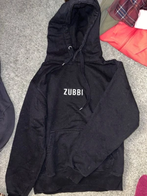 Svart hoodie ZUBBI från Dunderkläder LIMITED EDITION  - Svart hoodie från Dunderkläder med trycket 'ZUBBI' på bröstet. Klassisk modell med stor känguruficka framtill, justerbar dragsko i huvan och mjukt material. Perfekt för dig som gillar enkel och stilren streetwear.
