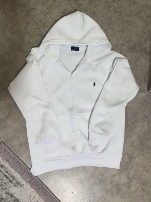 Vit hoodie från Polo Ralph Lauren - Säljer en clean vit hoodie från Polo Ralph Lauren med dragkedja och huva. Klassisk blå logga broderad på bröstet. Hoodien har fickor framtill och ribbade muddar vid ärmar och nederkant. Perfekt för en chill och stilren look.