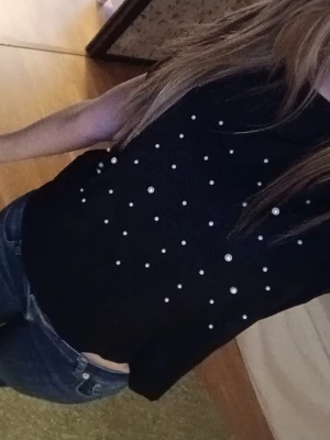 Svart topp med pärldetaljer - Svart kortärmad topp dekorerad med vita pärlor framtill. Modellen är rak och något croppad, vilket ger en trendig look. Perfekt att styla med jeans eller kjol för en cool outfit.