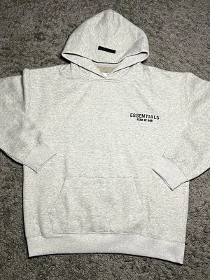 Grå Essentials Fear of God hoodie - Snygg ljusgrå hoodie från Essentials Fear of God med svart logga tryckt på bröstet och ryggen. Hoodien har en huva, ribbade muddar och en klassisk magficka. Tillverkad i mjukt bomullsmaterial som känns riktigt skönt mot huden.