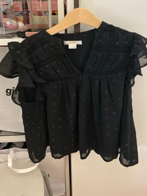 Svart blus med volanger från Gina Tricot - Snygg svart blus från Gina Tricot med volangdetaljer på axlarna och prickig struktur i tyget.  Har använts 1 gång‼️