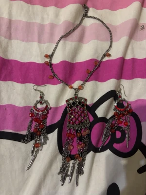 Bohemiskt smyckeset i silver och rött - Unikt smyckeset med halsband och matchande örhängen i silverfärgad metall. Dekorerade med röda och orange pärlor samt hängande kedjor och detaljer. Perfekt för dig som gillar bohemisk och färgstark stil. 