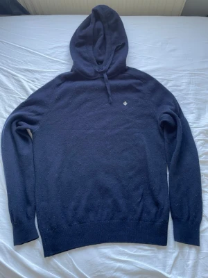 Mörkblå stickad hoodie från Morris - Mörkblå hoodie i mjukt stickat material med huva. Liten broderad logga på bröstet. Långa ärmar och ribbade muddar vid ärmslut och nederkant. Perfekt för en avslappnad och stilren look. Storlek L, den är något nopprig men annars i väldigt bra skick. Pris går att diskuteras