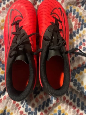  fotbollsskor - Säljer ett par Nike Phantom GT fotbollsskor i rött och svart med vita detaljer. Skorna har snörning, låg profil och syntetiskt material för bra bollkontroll. Swoosh-logga på sidan och Phantom GT-tryck bak. Perfekta för dig som vill sticka ut på planen.