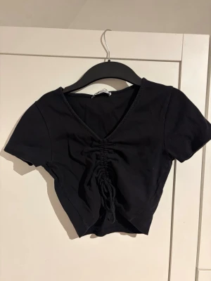 Svart croppad topp med resårband  - Trendig svart croppad topp med korta ärmar och djup V-ringning som går att ändra. Toppen har en snygg resårband framtill som ger en rynkad effekt och gör att längden kan justeras. Tillverkad i ribbad bomullsblandning för en bekväm och stretchig passform.