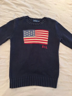Mörkblå stickad tröja Polo Ralph Lauren - Mörkblå stickad tröja från Polo Ralph Lauren med amerikansk flagga i rött, vitt och blått på bröstet samt RL-broderi. Tröjan har rund halsringning och ribbstickade muddar vid ärmslut och nederkant. Perfekt för dig som gillar klassisk preppy stil.