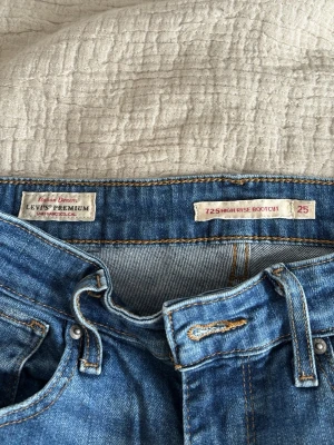 Levi's 725 High Rise Bootcut Jeans - Säljer ett par klassiska blå Levi's Premium 725 High Rise Bootcut jeans. De har hög midja och bootcut-ben, vilket ger en snygg siluett. Jeansen är i ett mjukt denimtyg med traditionella detaljer som fem fickor och bälteshällor.