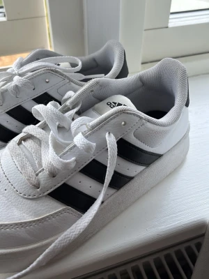 Vita Adidas sneakers med svarta ränder - Snygga vita sneakers från Adidas med de klassiska tre svarta ränderna på sidorna. Skorna har lågt skaft, vit sula och är tillverkade i läderliknande material med meshfoder. Perfekta för en clean och sportig stil.