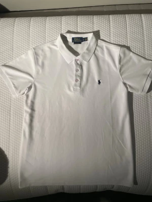 Vit pikétröja från Polo Ralph Lauren - Klassisk vit pikétröja från Polo Ralph Lauren med korta ärmar och knappar framtill. Tröjan har den ikoniska broderade loggan på bröstet och är tillverkad i mjuk bomull. Perfekt till en clean och stilren look.
