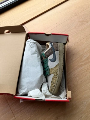 Nike Dunk Low Safari Golden Moss - Nike Dunk Low sneakers med djurmönster i brunt, grönt och beige. Skorna har gröna skosnören, beige meshpaneler och en mörkgrå swoosh. Yttersulan är beige och ovandelen har flera olika texturer och material för en unik look.