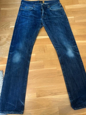 Blå raka jeans från Nudie Jeans - Säljer ett par klassiska blå jeans från J.CREW med rak passform och fem fickor. Jeansen har snygga slitningar vid knäna och bak, samt kontrastsömmar. Tillverkade i denim med en autentisk tvättad look. Perfekta för en avslappnad stil. SELVEGE JEANS 