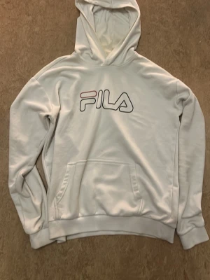 Vit hoodie från FILA med logga - Snygg vit hoodie från FILA med stor logga i svart och rött på bröstet. Klassisk känguruficka framtill och ribbade muddar vid ärmslut och nederkant. Tillverkad i mjukt material som känns skönt mot huden. Perfekt för en avslappnad och sportig stil. Små defekter, inga hål. Kolla storlek på bilden! Det mesta är damm, skriv gärna vid funderingar!